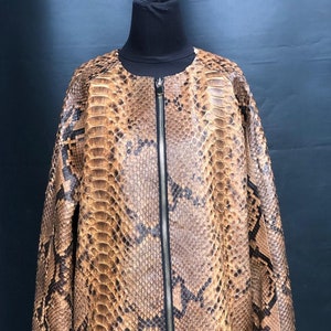 以下が含まれることがあります： 茶色と黒のヘビ革ジャケット、フルレングスのジッパー付き。 ジャケットは黒いマネキンに展示されています。 ヘビ革の模様がジャケット全体に施されており、茶色と黒の鱗が混ざっています。