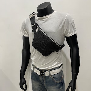 May include: A black leather fanny pack with a silver zipper and buckle. The fanny pack is worn on a mannequin over a white t-shirt and blue jeans.