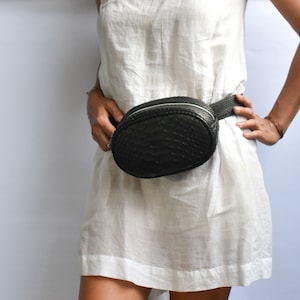 May include: A black leather fanny pack with a snakeskin pattern. The pack is worn around the waist of a person wearing a white dress.