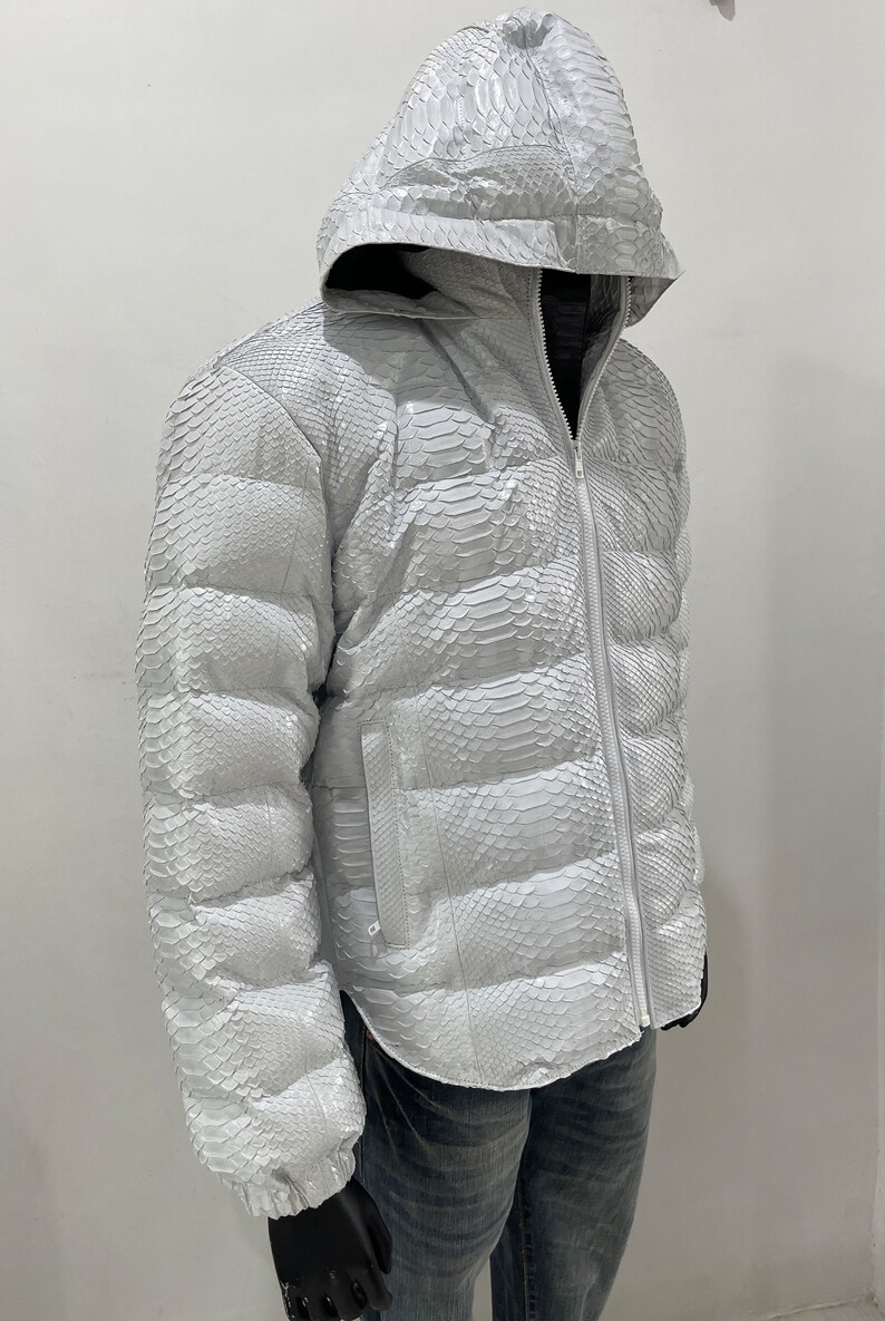 May include: A white puffer jacket made of snakeskin with a hood. The jacket has a zip closure and a quilted design.