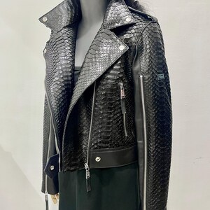 K&ouml;nnte beinhalten: Schwarze Lederjacke mit Schlangenhautmuster. Die Jacke hat einen Rei&szlig;verschluss und einen Stehkragen.