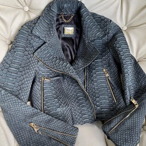 以下が含まれることがあります： 金色のジッパーと金色のチェーンのディテールが付いたグレーのヘビ革のバイクジャケット。ジャケットの前面には「DEER」のラベルが付いています。