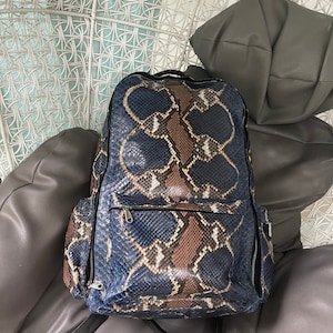 May include: A blue and brown snakeskin backpack with two zipped pockets on the sides. The backpack is sitting on a white wicker chair.