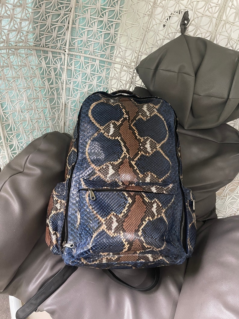 Op de afbeelding: Een blauwe en bruine slangleren rugzak met twee ritszakken aan de zijkanten. De rugzak staat op een witte rieten stoel.