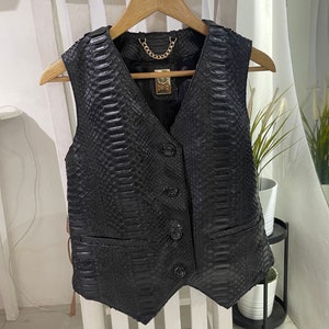 May include: A black leather vest with a snakeskin pattern and a gold chain detail. The vest has a V-neck and four buttons down the front.
