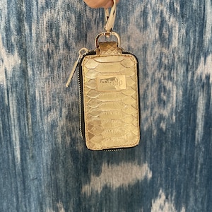 May include: Gold key chain with a black zippered pouch and a gold metal clasp. The pouch has a snakeskin pattern and the text "to help" is printed on the front.