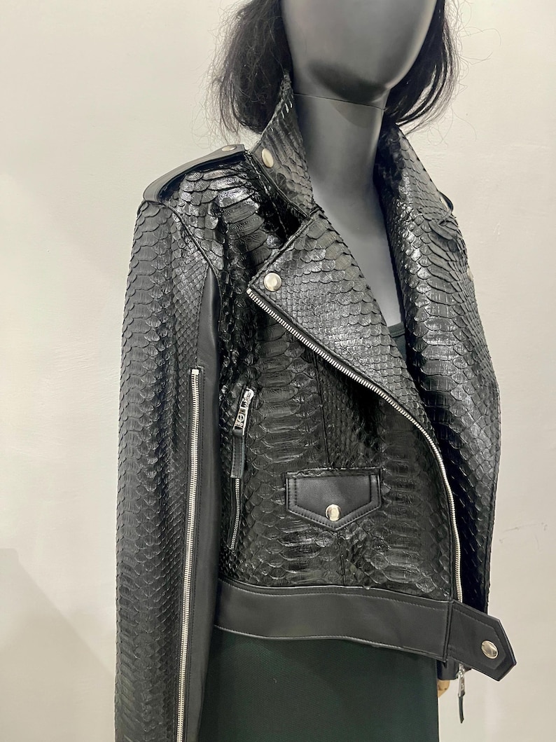 K&ouml;nnte beinhalten: Schwarze Lederjacke mit Schlangenhautmuster. Die Jacke hat einen Rei&szlig;verschluss, einen Kragen und zwei Taschen.