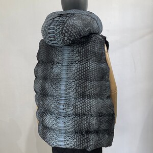 May include: A sleeveless puffer vest with a snakeskin pattern in shades of grey and blue. The vest has a hood and is quilted for warmth.