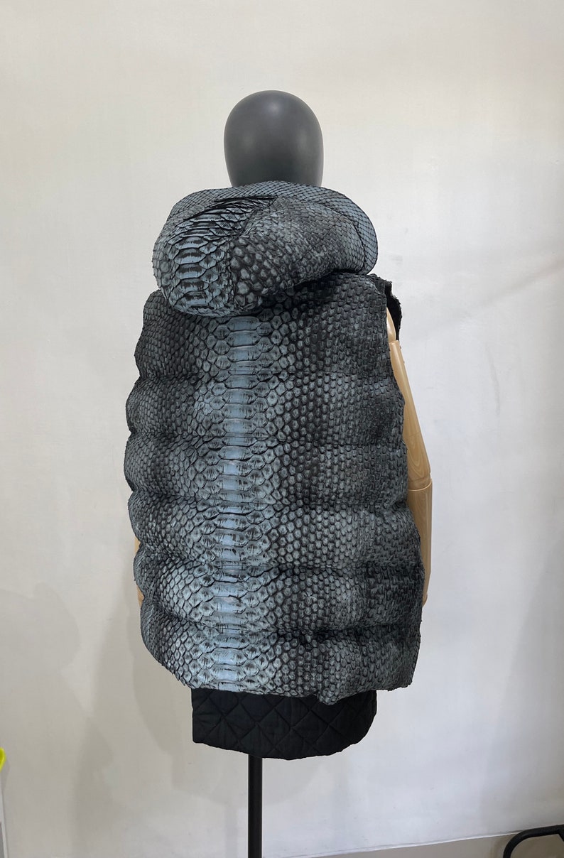 May include: A sleeveless puffer vest with a snakeskin pattern in shades of grey and blue. The vest has a hood and is quilted for warmth.