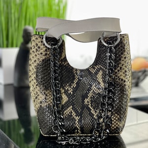 以下が含まれることがあります： グレーの革のストラップと黒いチェーンストラップが付いた、黒と茶色のヘビ柄の小さなハンドバッグです。