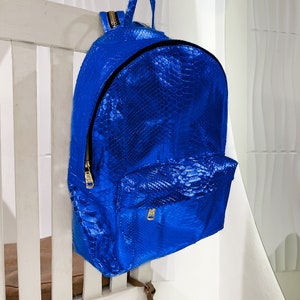 以下が含まれることがあります： 金色のジッパーと金色の引き手のある明るい青色のヘビ革のバックパック。バックパックは白い木製の椅子にかけられています。