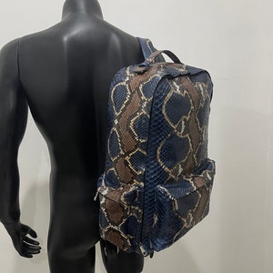 Op de afbeelding: Een blauwe en bruine slangenleren rugzak met meerdere vakken en een ritssluiting.