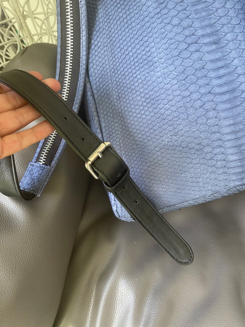 K&ouml;nnte beinhalten: Eine blaue Tasche aus Schlangenleder mit einem verstellbaren schwarzen Riemen und einem silbernen Rei&szlig;verschluss. Die Tasche liegt auf einer grauen, strukturierten Oberfl&auml;che.