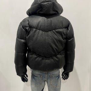 Op de afbeelding: Zwarte pufferjack met capuchon, met een slangenleerpatroon. De jas heeft een gewatteerd ontwerp en een trekkoord in de taille. Het model draagt een blauwe spijkerbroek en zwarte handschoenen.