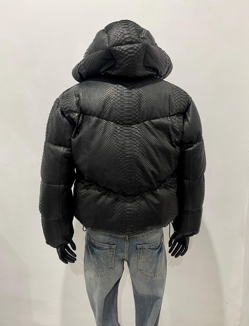 Op de afbeelding: Zwarte pufferjack met capuchon, met een slangenleerpatroon. De jas heeft een gewatteerd ontwerp en een trekkoord in de taille. Het model draagt een blauwe spijkerbroek en zwarte handschoenen.