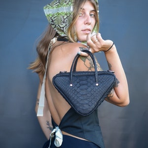 May include: A woman wearing a black top and pants with a snakeskin print headscarf and a black triangular handbag with a snakeskin pattern.