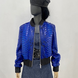 以下が含まれることがあります： 黒いジッパーと黒いリブトリムが付いた青いヘビ革のボンバージャケット。ジャケットは、黒いベレー帽とデニムスカートを着たマネキンに着せられています。