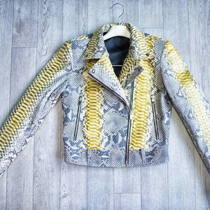 以下が含まれることがあります： グレー、イエロー、ホワイトの色合いのヘビ柄のスタイリッシュなジャケット。ノッチカラー、非対称ジッパー、ジッパーポケットを備えたバイカースタイルのジャケットです。白いハンガーにかかっています。