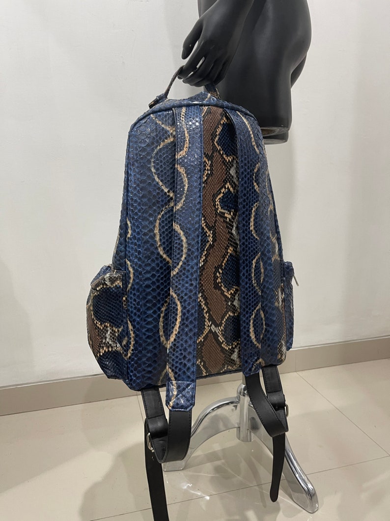 Op de afbeelding: Een blauwe en bruine slangenleren rugzak met zwarte banden. De rugzak heeft een ritsvak aan de voorkant en een bovenste handvat.