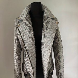 以下が含まれることがあります： ジッパー留めとベルトが付いたグレーのヘビ革ジャケット。ジャケットはスタンドカラーと長袖です。