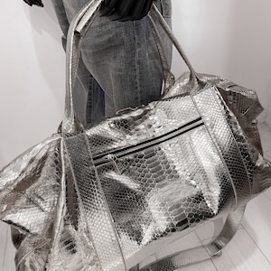 May include: A silver snakeskin leather duffel bag with a zipper closure and a long shoulder strap.