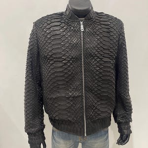 以下が含まれることがあります： 黒い革のジャケット、ヘビ革模様。 ジャケットはジッパー開閉、リブ編みの襟、リブ編みの袖口が付いています。 ジャケットは、ジーンズを履いたマネキンに展示されています。