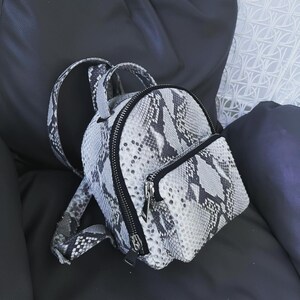 May include: A small, white and black snakeskin patterned backpack with black zipper accents and a black adjustable strap.