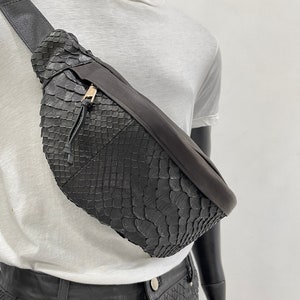 以下が含まれることがあります： ヘビ柄の黒い革製のウエストバッグ。バッグには金色のジッパーと黒いストラップが付いています。