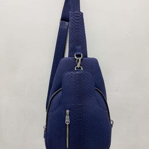 以下が含まれることがあります： ネイビーブルーのスリングバッグ。表面は蛇革風のテクスチャ加工。調節可能なストラップ1本、ジッパー付きフロントポケット、シルバーカラーの金具付き。洗練されたモダンなデザインで、普段使いに適しています。