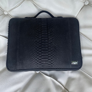May include: A black leather laptop case with a snakeskin pattern and a small logo on the bottom right corner.