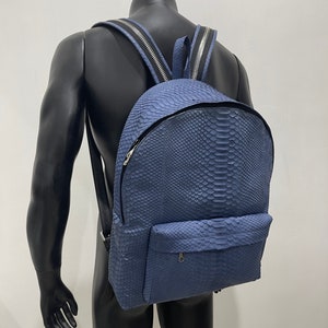 K&ouml;nnte beinhalten: Ein blauer Lederrucksack mit Schlangenhautmuster. Der Rucksack hat eine Rei&szlig;verschlusstasche vorne und zwei verstellbare Tr&auml;ger.