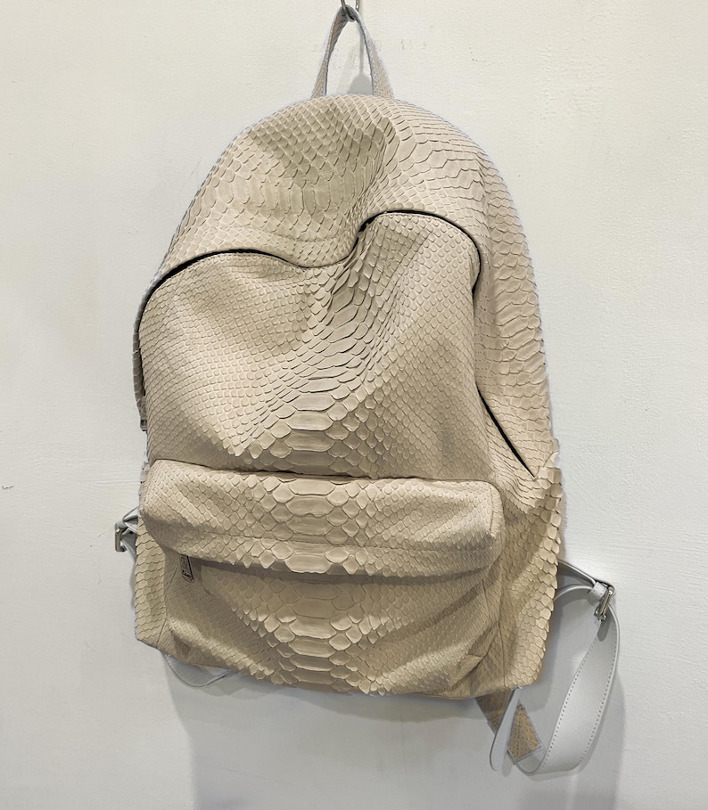 Op de afbeelding: Een beige rugzak van slangenleer met een verstelbare witte band. De rugzak heeft een ritsvak aan de voorkant.