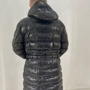 May include: A long, black puffer coat with a hood. The coat has a textured, snakeskin-like pattern and a cinched waist. The coat is designed to be worn in cold weather.