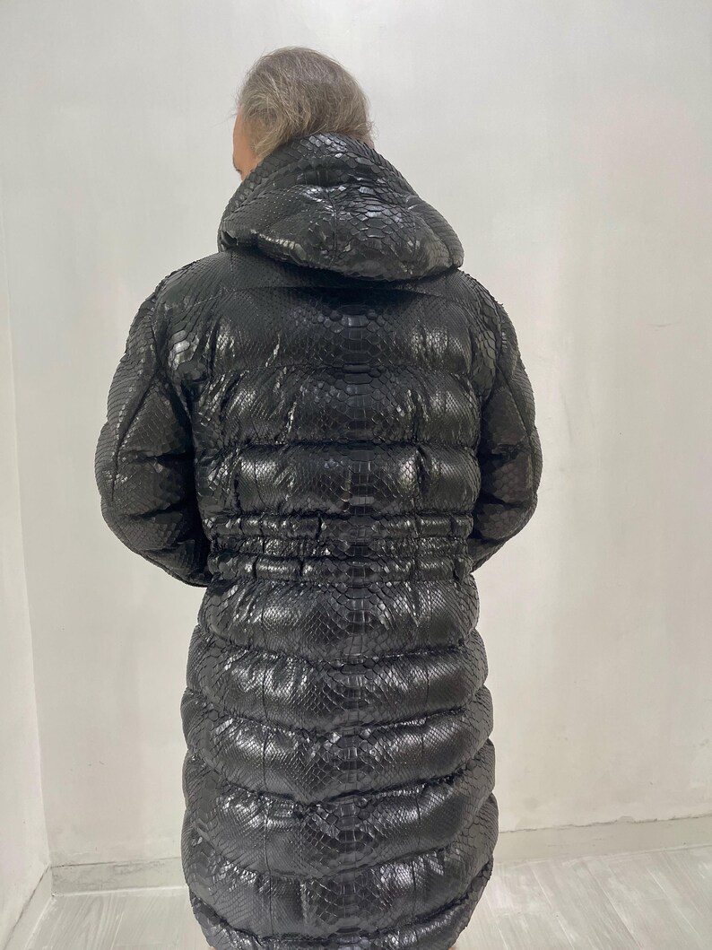 May include: A long, black puffer coat with a hood. The coat has a textured, snakeskin-like pattern and a cinched waist. The coat is designed to be worn in cold weather.