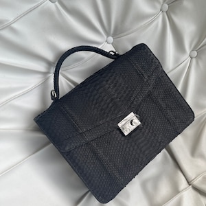 May include: A black snakeskin-patterned handbag with a silver clasp and a top handle. The rectangular bag features a flap closure and is set against a white, button-tufted background.