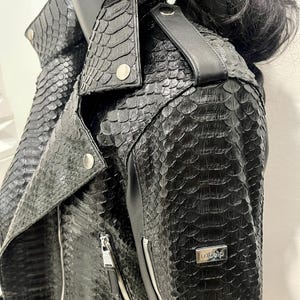 K&ouml;nnte beinhalten: Schwarze Lederjacke mit Schlangenhautmuster und silbernen Rei&szlig;verschlussdetails. Die Jacke hat einen Kragen und einen Rei&szlig;verschluss. Der Markenname "O.elp" ist auf der rechten Seite der Jacke sichtbar.