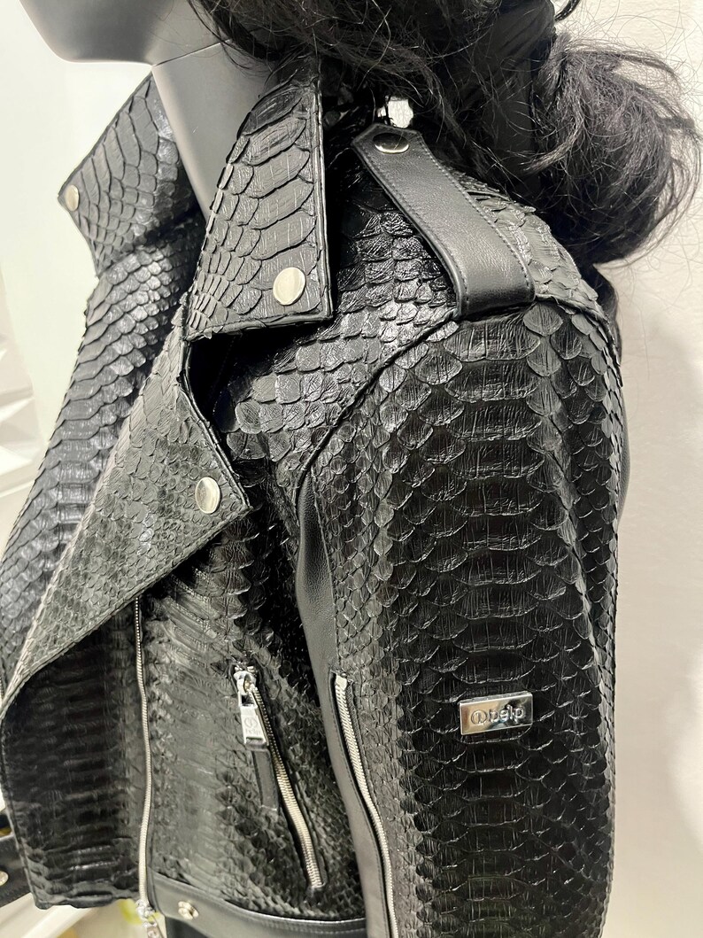 K&ouml;nnte beinhalten: Schwarze Lederjacke mit Schlangenhautmuster und silbernen Rei&szlig;verschlussdetails. Die Jacke hat einen Kragen und einen Rei&szlig;verschluss. Der Markenname "O.elp" ist auf der rechten Seite der Jacke sichtbar.