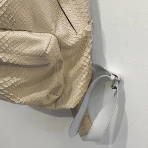 Op de afbeelding: Een lichtbeige rugzak van slangenleer met een witte leren riem en een zilveren gesp.