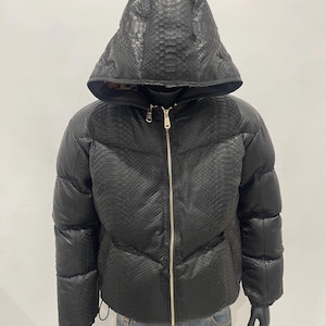 以下が含まれることがあります： スネークスキン柄のフード付き黒いパファージャケット。 ジャケットはゴールドのジッパーが付いており、マネキンに展示されています。 ジャケットは冬用のアウターウェアです。