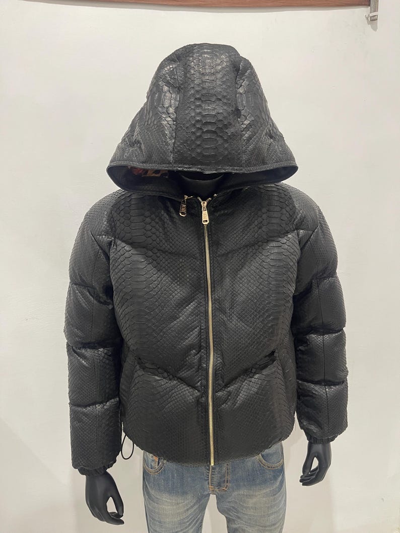 Op de afbeelding: Een zwarte pufferjack met capuchon, met een slangenleerpatroon. De jas heeft een gouden rits en wordt op een mannequin getoond. De jas is een winterjas.