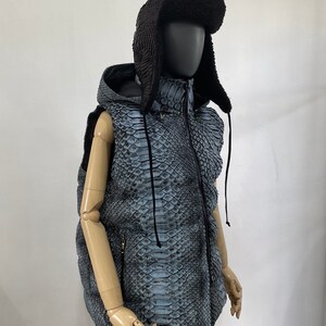 May include: A blue and black hooded vest with a snakeskin pattern. The vest has a zipper closure and a drawstring hood. The vest is being modelled on a mannequin wearing a black fur hat.