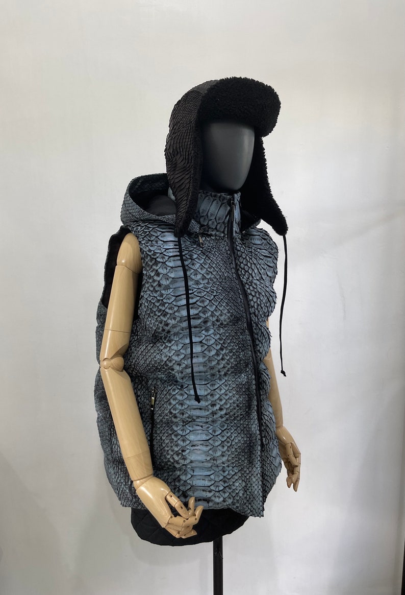 May include: A blue and black hooded vest with a snakeskin pattern. The vest has a zipper closure and a drawstring hood. The vest is being modelled on a mannequin wearing a black fur hat.