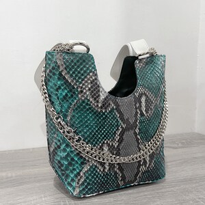 以下が含まれることがあります： 緑と灰色のヘビ革のハンドバッグに銀色のチェーンストラップが付いている。