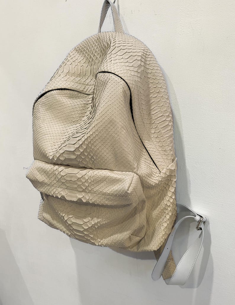 Op de afbeelding: Een lichtbeige rugzak van slangenleer met een witte leren band. De rugzak heeft een ritsvak aan de voorkant en een bovenste handvat.