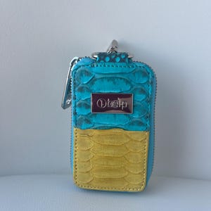 May include: A rectangular, zipped key case with a teal and yellow snakeskin pattern. The top half is teal, the bottom half is yellow. A silver metal plate with the word "Ohelp" is centered on the teal section. A silver metal clasp is attached.