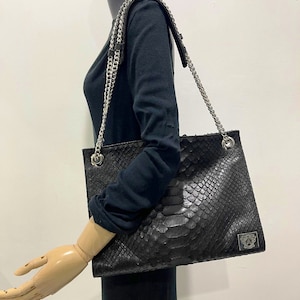 以下が含まれることがあります： 蛇柄のブラックレザーのトートバッグ。シルバーのチェーンストラップが付いています。バッグの右下にはシルバーのロゴがあります。