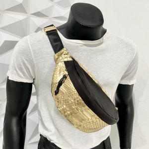 May include: A gold snakeskin fanny pack with a black nylon strap and a black nylon flap. The bag is worn on a mannequin.