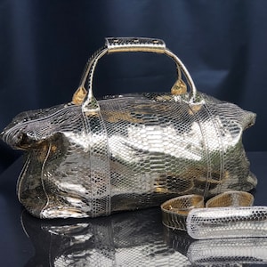 May include: A gold-coloured snakeskin leather duffel bag with a top handle and a matching strap. The bag is lying on a black surface.