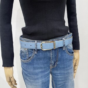 May include: A light blue leather belt with a gold buckle. The belt is worn over a pair of blue jeans.