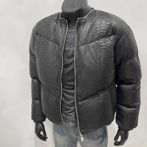 Op de afbeelding: Zwarte pufferjack met slangenpatroon, ritssluiting en ronde kraag. Gepresenteerd op een mannequin, gecombineerd met een grijs shirt en een blauwe jeans. Het ontwerp van de jas omvat een gewatteerd patroon.